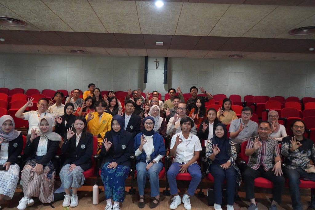 A group photo of about 30 participants, including Ecoton members and UKWMS students and faculty, seated in red auditorium chairs. Everyone is smiling and holding up an "OK" hand gesture. Prigi Arisandi and Dr. Herjunianto are visible in the front row among the participants.