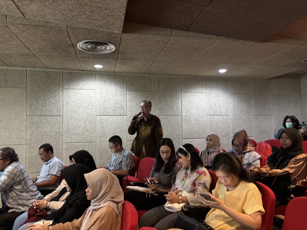 Dr. Roy Hendroko Setyobudi, M.Si, wearing a patterned batik shirt, stands at a podium giving a presentation on "Pencegahan Bahaya Kesehatan" (Health Hazard Prevention). He is addressing a panel and audience at UKWMS Surabaya during an Ecoton Focus Group Discussion.