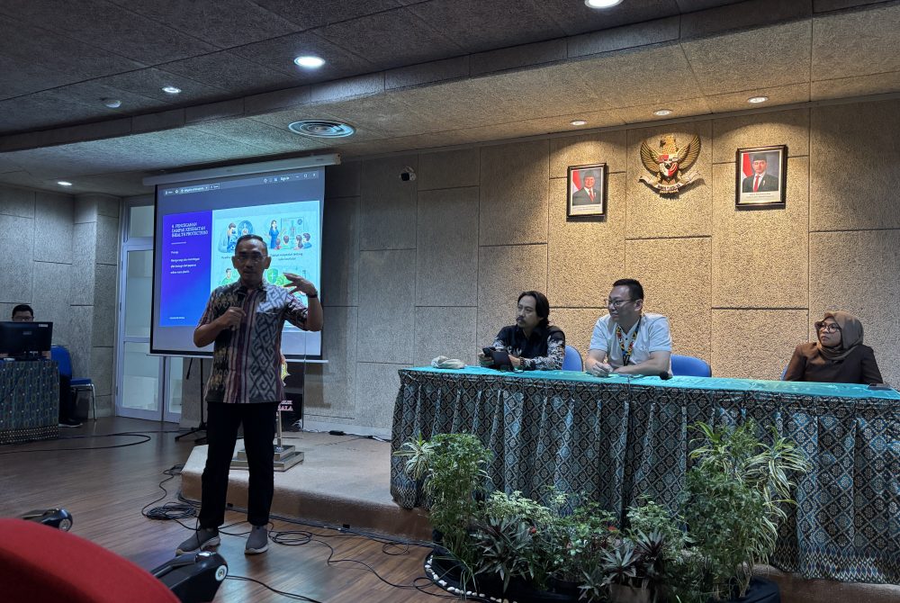 Laksamana Pertama TNI Purn. Dr. Herjunianto SpPD, MMRS, the Dean of the Faculty of Medicine at UKWMS, stands at the front of a hall speaking into a microphone. Behind him is a presentation slide titled "Pencegahan Bahaya Kesehatan" (Health Hazard Prevention). Three panelists sit at a draped table to his right in a formal setting featuring the Indonesian national emblem.