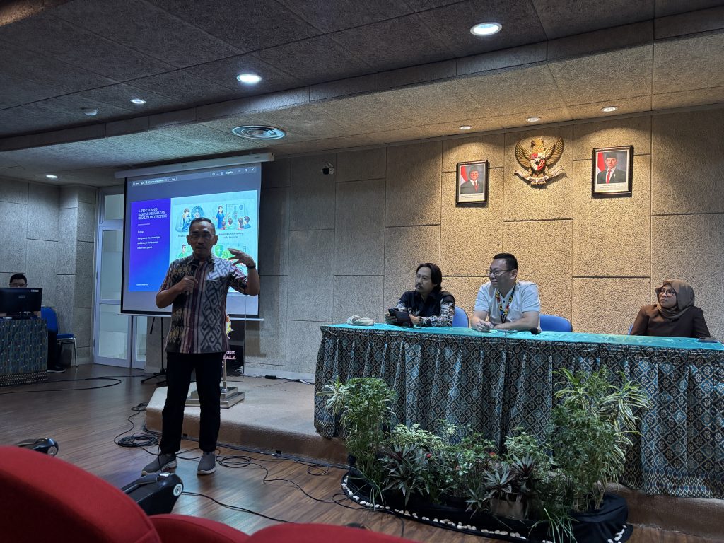 Laksamana Pertama TNI Purn. Dr. Herjunianto SpPD, MMRS, the Dean of the Faculty of Medicine at UKWMS, stands at the front of a hall speaking into a microphone. Behind him is a presentation slide titled "Pencegahan Bahaya Kesehatan" (Health Hazard Prevention). Three panelists sit at a draped table to his right in a formal setting featuring the Indonesian national emblem.