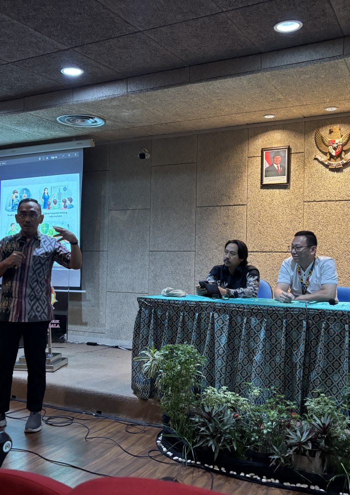 Laksamana Pertama TNI Purn. Dr. Herjunianto SpPD, MMRS, the Dean of the Faculty of Medicine at UKWMS, stands at the front of a hall speaking into a microphone. Behind him is a presentation slide titled "Pencegahan Bahaya Kesehatan" (Health Hazard Prevention). Three panelists sit at a draped table to his right in a formal setting featuring the Indonesian national emblem.