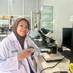 A female researcher wearing a white lab coat and a brown hijab sits at a laboratory desk with a microscope and Petri dish.