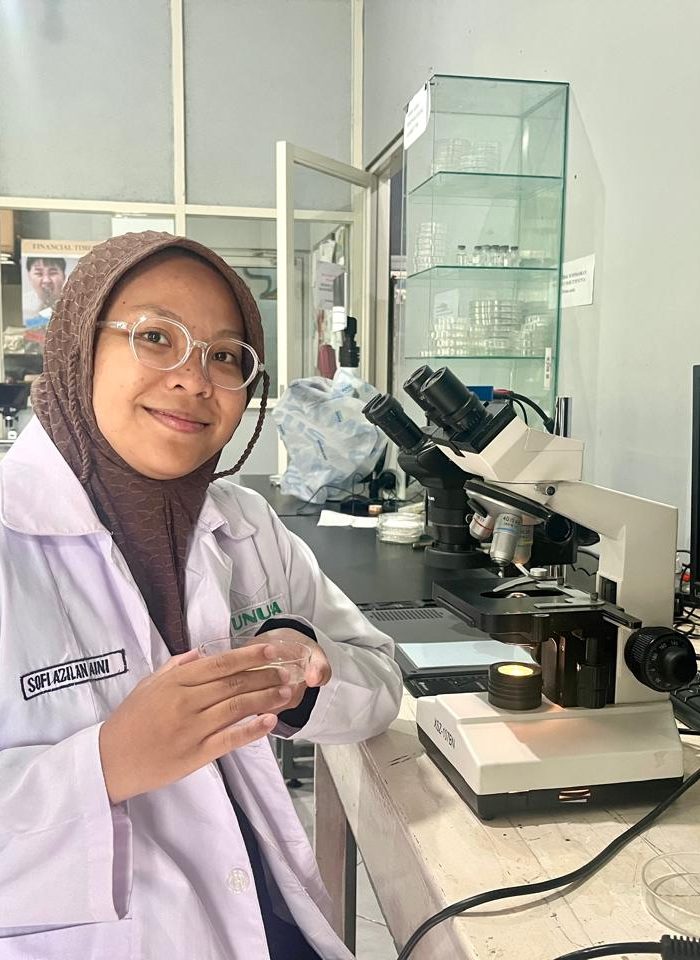 A female researcher wearing a white lab coat and a brown hijab sits at a laboratory desk with a microscope and Petri dish.