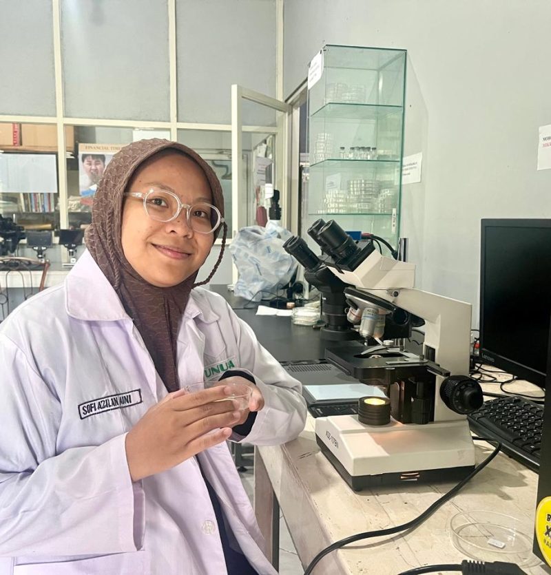 A female researcher wearing a white lab coat and a brown hijab sits at a laboratory desk with a microscope and Petri dish.