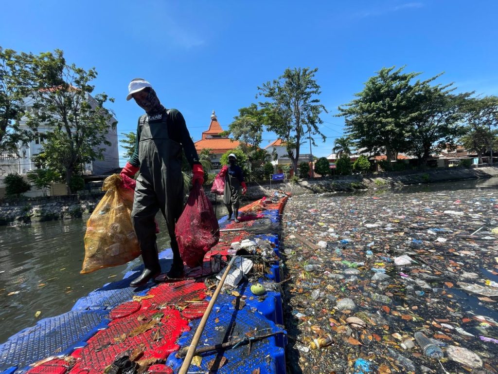 Volunteers carry sacks of collected plastic waste while cleaning Kali Tebu River using a floating platform.