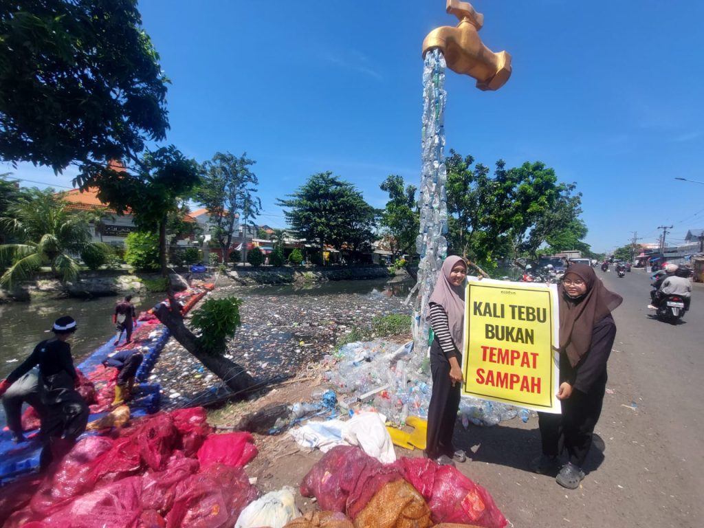 Volunteers clean piles of plastic waste in Kali Tebu River using nets and a floating platform, with a giant faucet installation made from plastic bottles and a “Kali Tebu is Not a Dumping Site” sign on the riverbank.