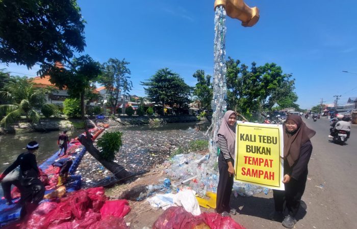 Volunteers clean piles of plastic waste in Kali Tebu River using nets and a floating platform, with a giant faucet installation made from plastic bottles and a “Kali Tebu is Not a Dumping Site” sign on the riverbank.