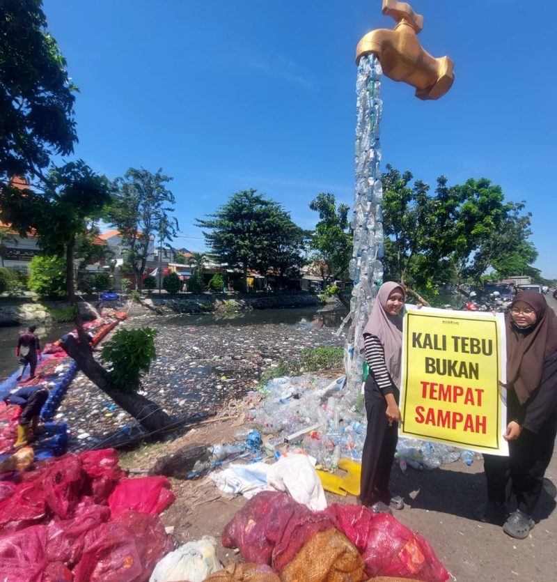 Volunteers clean piles of plastic waste in Kali Tebu River using nets and a floating platform, with a giant faucet installation made from plastic bottles and a “Kali Tebu is Not a Dumping Site” sign on the riverbank.